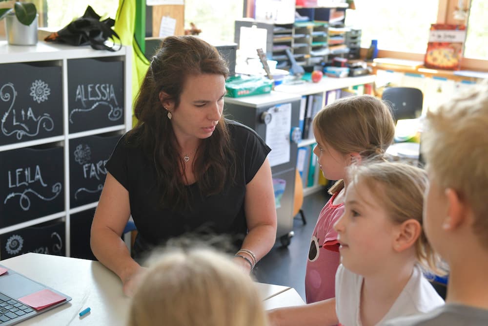 Eine Frau im Gespräch mit vier Kindern. Die vier Kinder sind von der Seite und hinten abgebildet, die Frau frontal. Im Hintergrund ein Schulzimmer.