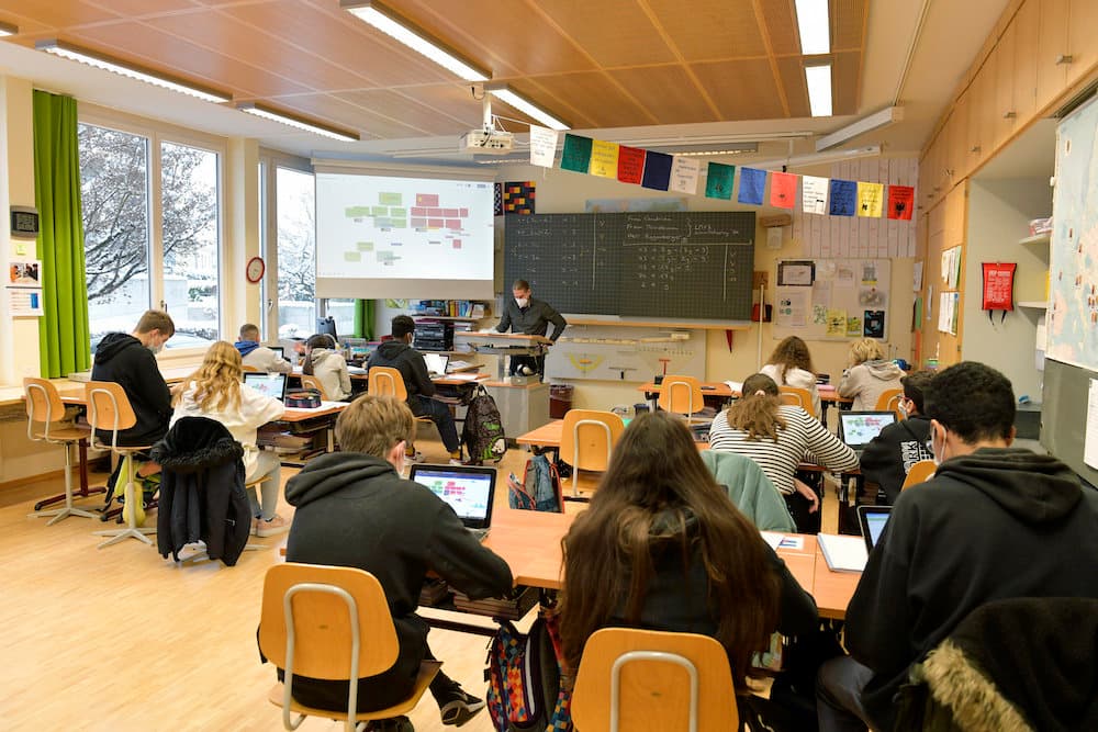 Ein Schulzimmer. Eine Gruppe von Jugendlichen sitzt an Schulpulten, vorne steht ein Lehrer vor der Wandtafel.