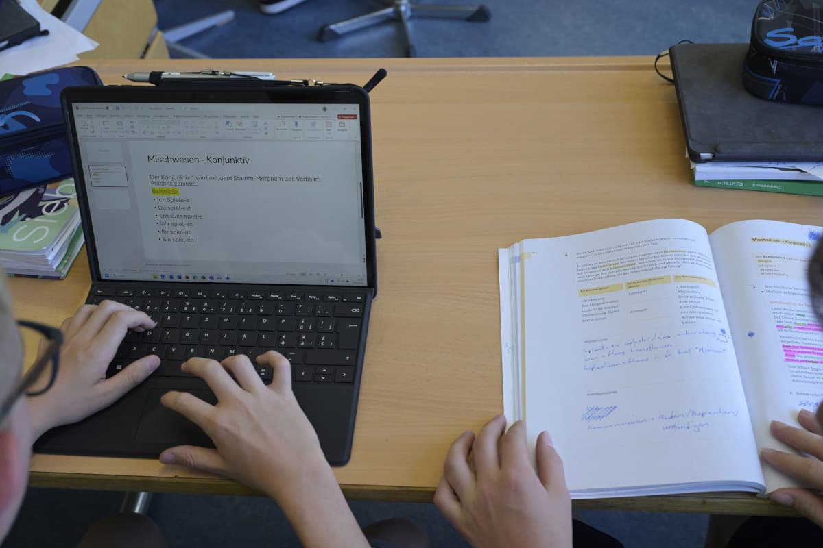 Ein Laptop und ein aufgeschlagenes Schulbuch liegen auf einem Tisch. Die Hände von zwei Jugendlichen sind sichtbar, sie arbeiten auf der Tastatur und halten das Buch.