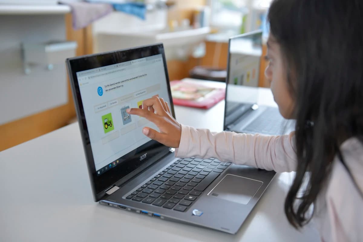 Ein Laptop auf einem Tisch auf der linken Seite, ein Mädchen tippt mit dem Finger auf den Bildschirm. Das Mädchen ist verschwommen von der Seite zu sehen, sie hat schwarze, lange Haare.
