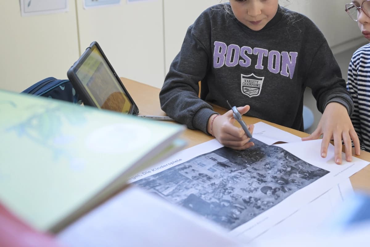 Zwei Kinder, ihre Gesichter abgeschnitten, arbeiten an einem Tisch, wo Schere, Bücher, Tablet und Arbeitsblätter zu sehen sind.