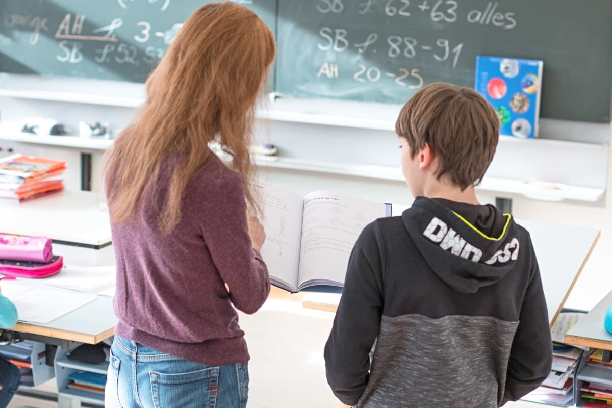 Eine erwachsene Person und ein Kind stehen vor einer Wandtafel, auf der Zahlen stehen. Sie schauen in ein geöffnetes Buch, dass die Frau mit langen roten Haaren in den Händen hält.