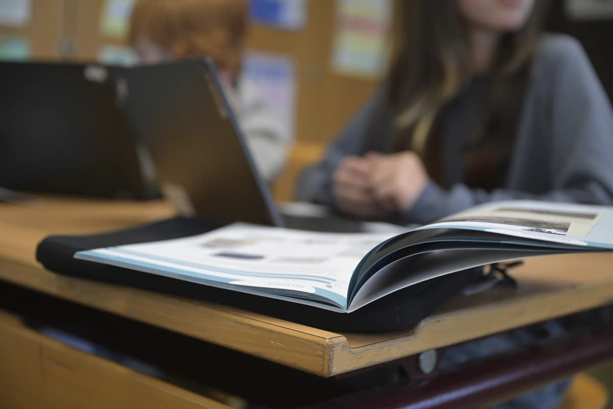 Ein offenes Arbeitsheft im Vordergrund auf einem Tisch, im Hintergrund verschwommen ein Kind am Laptop.