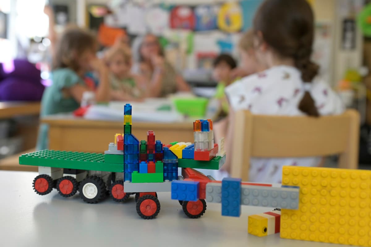 Im Vordergrund ein aus Lego gebautes Fahrzeug, im Hintergrund unscharf eine Gruppe von Kindern, die an einem Tisch sitzen.