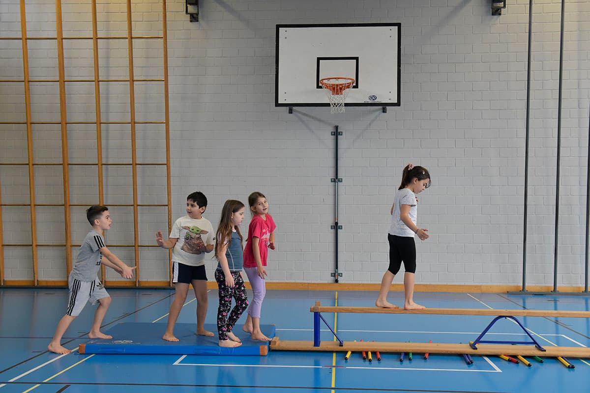 Ein Kind balanciert über eine Bank in der Turnhalle, andere Kinder reden.