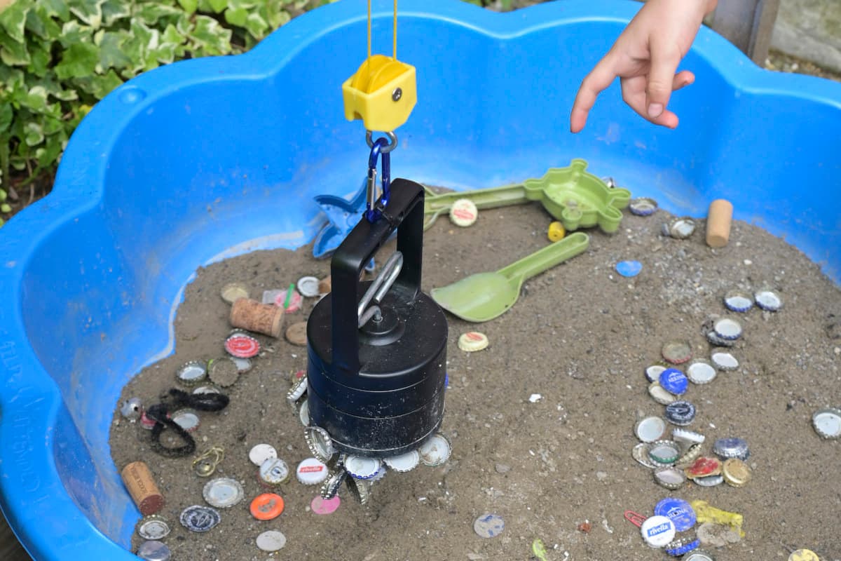 Ein Sandkasten gefüllt mit Sand und magnetischen Flaschendeckeln. Über dem Sandkasten hängt eine magnetische Winde, an ihr haften mehrere Flaschendeckel.