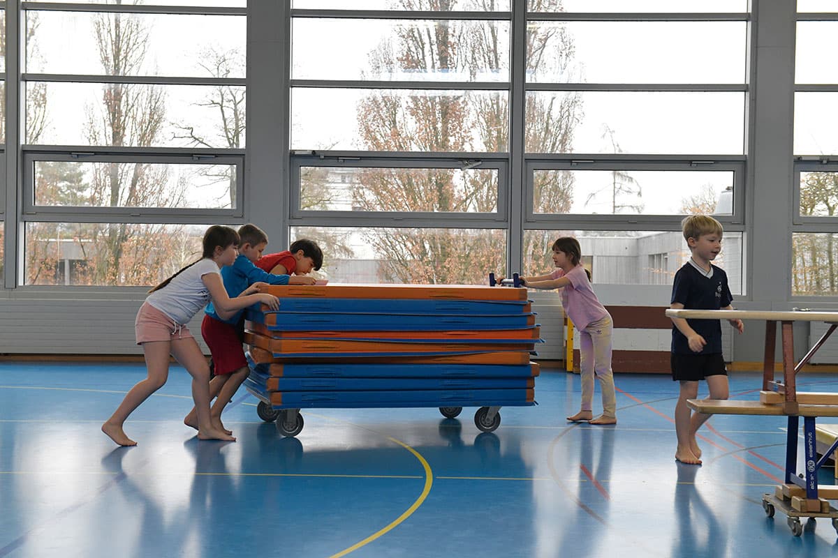 Kinder schieben einen Mattenwagen durch die Turnhalle.