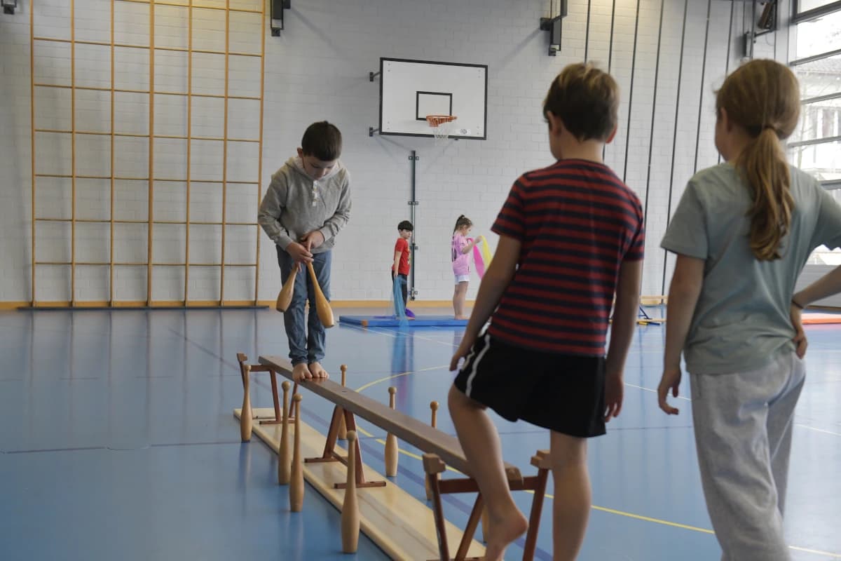 Drei Kinder balancieren auf einer Bank. Sie befinden sich in einer Turnhalle, im Hintergrund sind zwei weitere Kinder zu sehen.
