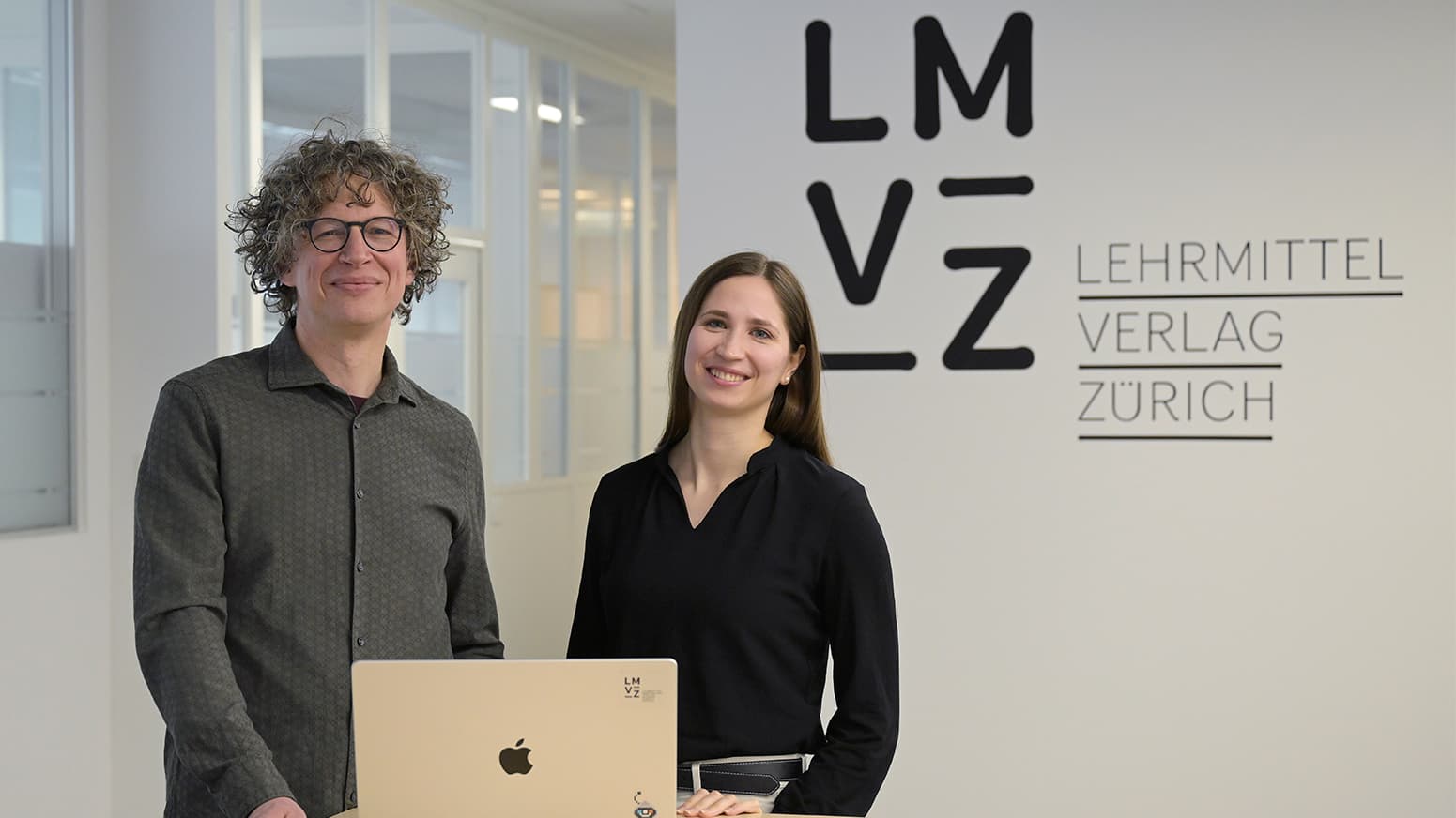 Two people smiling in an office with a laptop, standing in front of a wall displaying "Lehrmittel Verlag Zürich" and "LMVZ" logo.