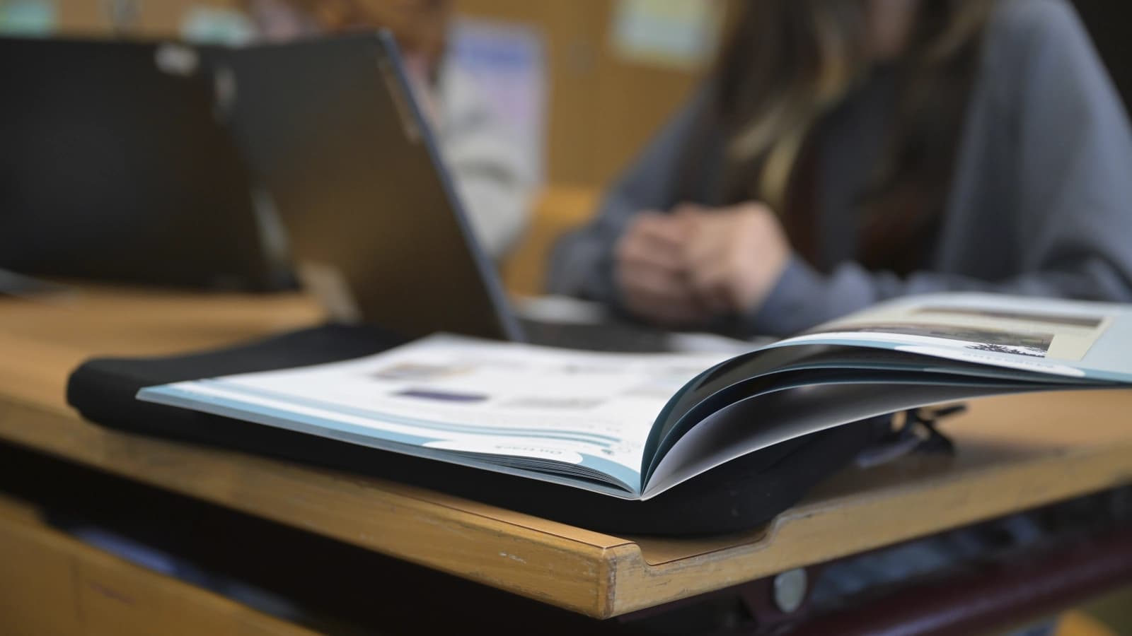 Ein offenes Heft und im Hintergrund ein Laptop und eine Person, von der man den Kopf nicht sieht.
