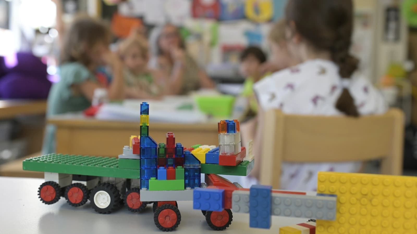 Im Vordergrund ein Spielzeug aus Lego, im Hintergrund verschwommen fünf Kinder und eine erwachsene Person, die um einen Tisch sitzen.