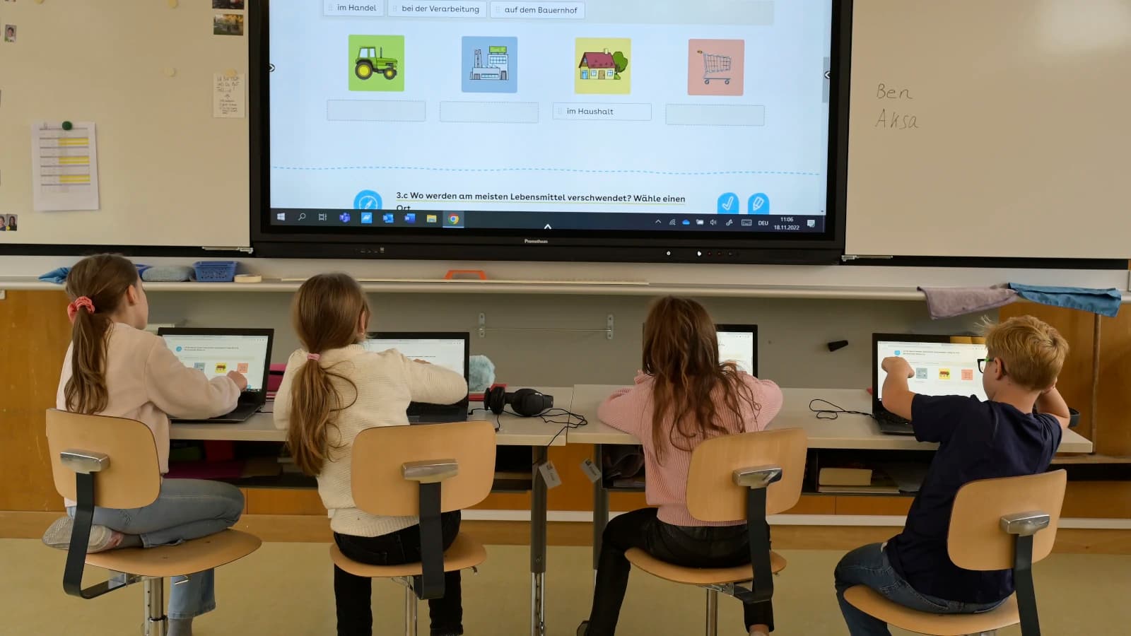 Vier Kinder sitzen in einer Reihe vor ihren Laptops, über ihnen wird auf einer Leinwand ein Bildschirminhalt eingeblendet. Die Kinder sind nur von hinten zu sehen, sie sitzen an einem langen Tisch.