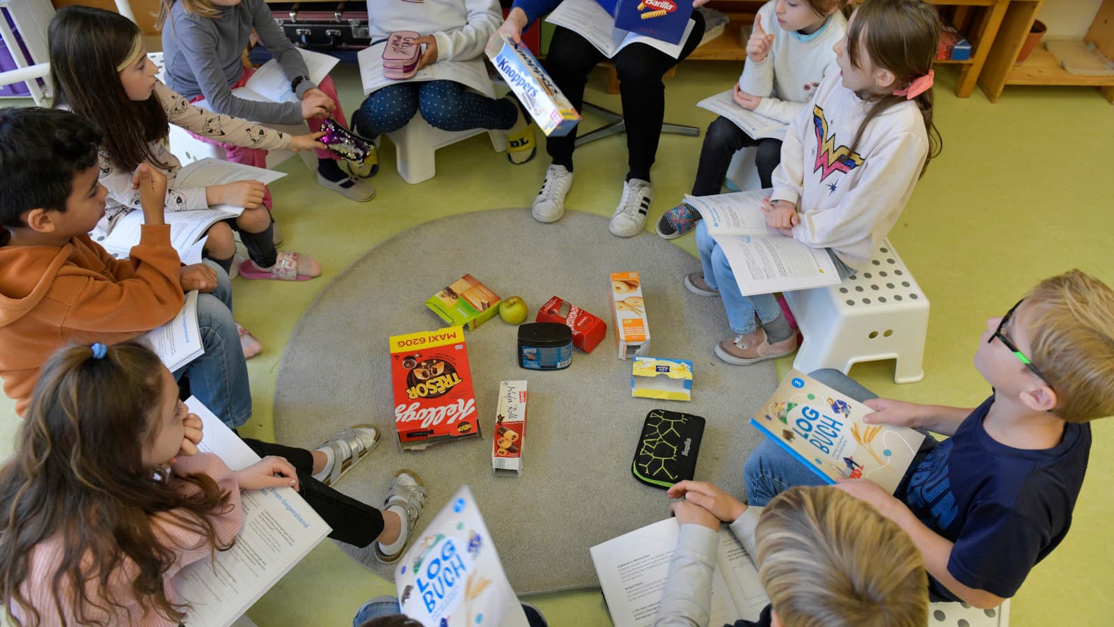 Kinder sitzen im Kreis, sie sind schräg von oben fotografiert. Sie betrachten verschiedene Gegenstände, die auf einem Teppich in der Mitte liegen.