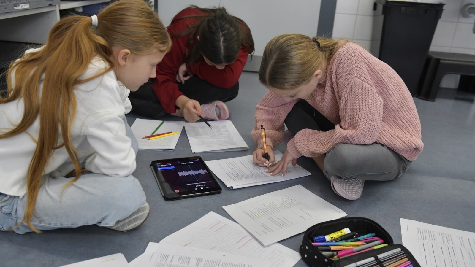 Drei Kinder, alle mit langen Haaren, sitzen am Boden und sind über Blätter und ein Tablett gebeugt.