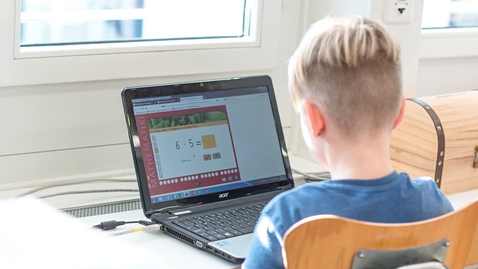 Ein etwa siebenjähriges, blondes Kind mit kurzen Haaren sitzt vor einem Computer und löst Matheaufgaben. Man sieht das Kind nur von hinten, auf dem Bildschirm ist die Aufgabe 6 mal 5 zu lesen.
