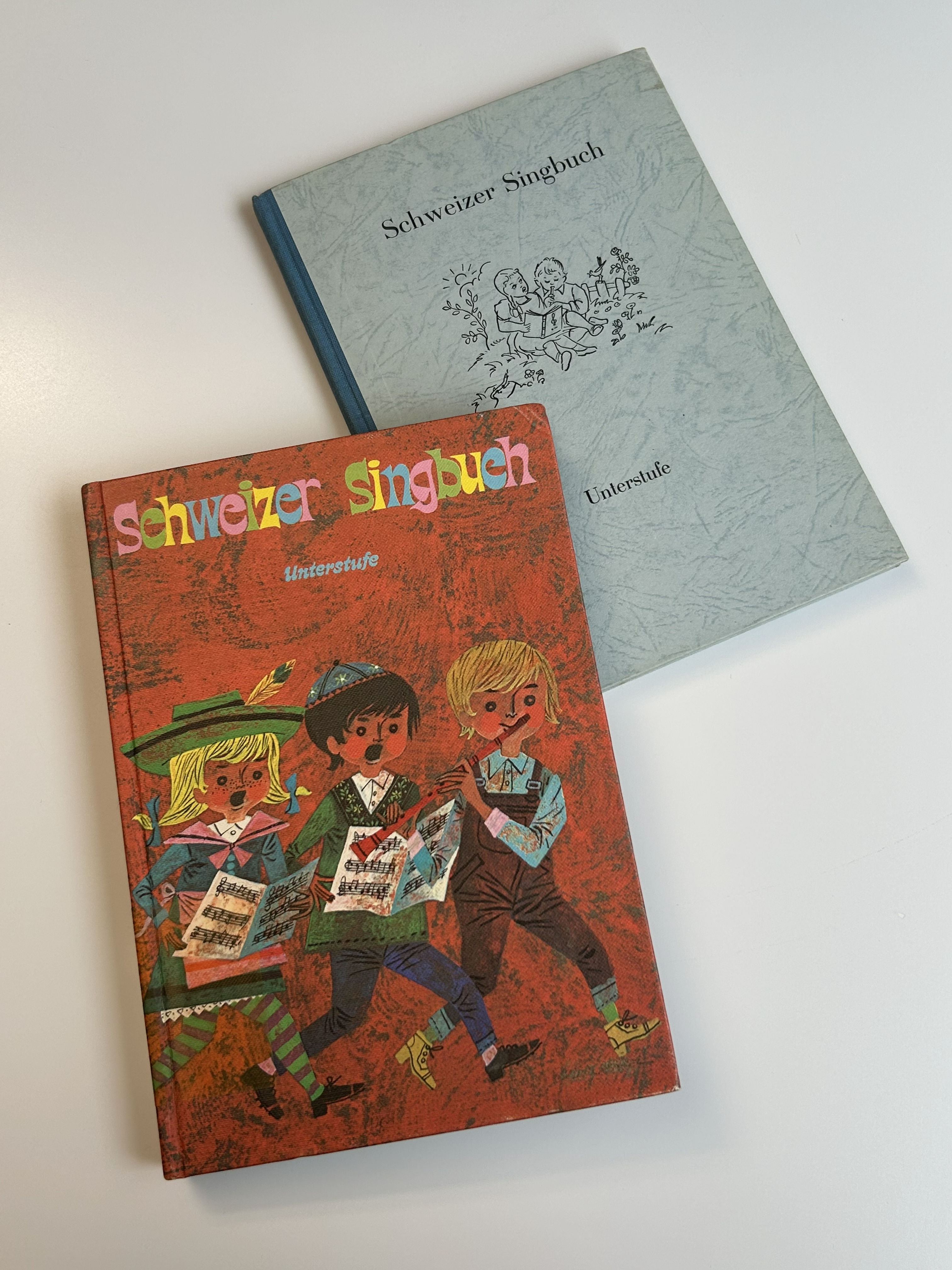 Das Cover vom Schweizer Singbuch für die Unterstufe ist zusehen. Ein blondes Mädchen, ein Junge mit schwarzem Haar und ein Junge mit blondem Haar, dass Flöte spielt.