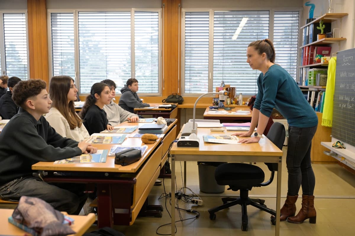 Ein Klassenzimmer, link sitzen Jugendliche in den Bänken und schauen nach vorne, wo eine Lehrerin am Pult steht.