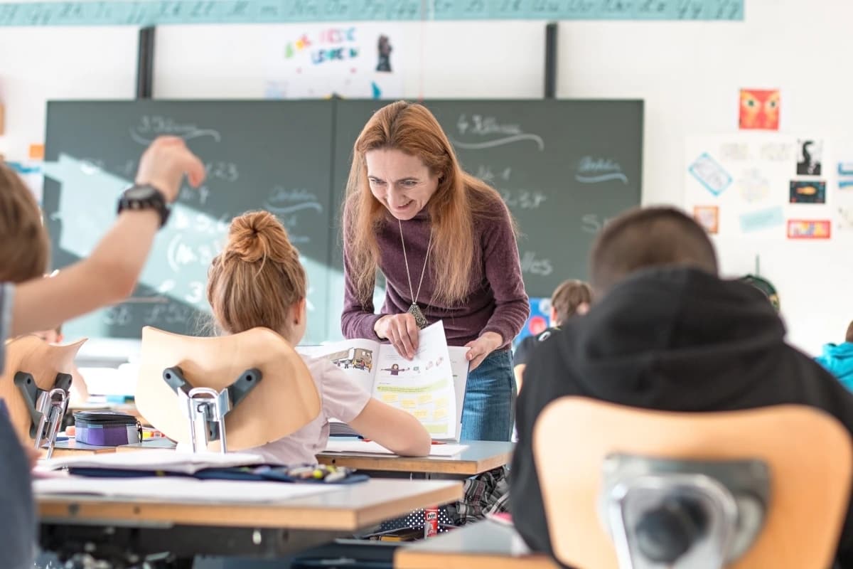 Ein Schulzimmer, die Kinder sind von hinten zu sehen. Eine Lehrerin mit langen roten Haaren beugt sich zu einem Kind hinunter und zeigt etwas im Buch.