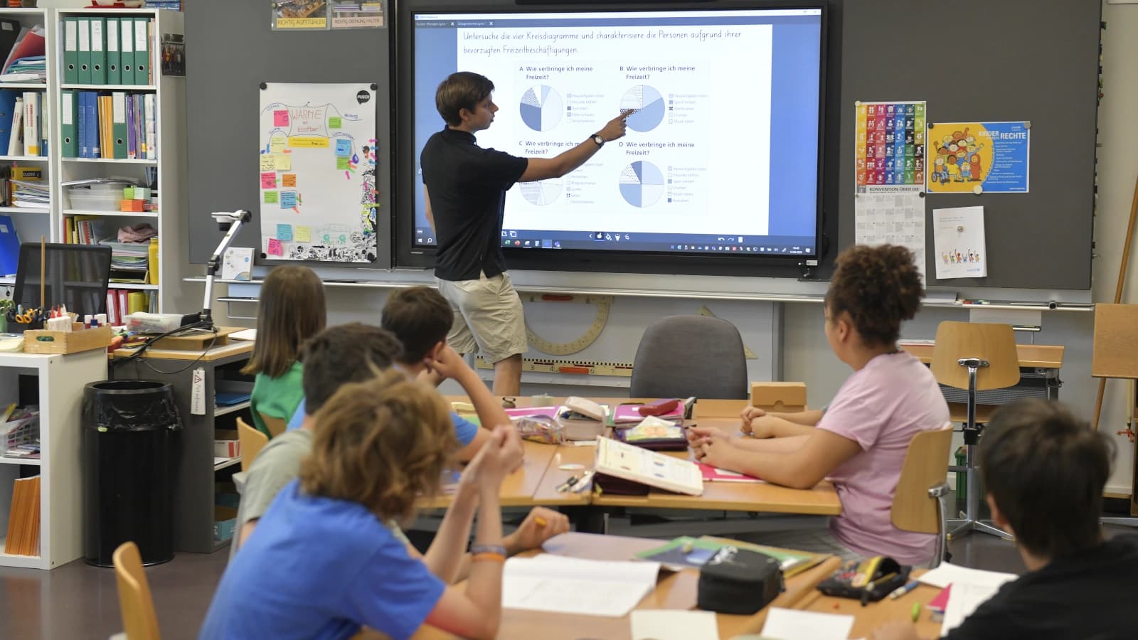 Ein Schulzimmer, an der Wandtafel steht ein Lehrer in schwarzem Shirt. Die Kinder sitzen an den Tische, sind nur von hinten zu sehen.